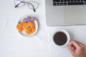 Hand holding coffee cup and eating Halloween Cookies during using computer laptop. Happy Halloween, online shopping, Hello October, fall autumn, Festive, party and holiday concept