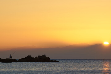 Obraz premium A man is fishing by the mediterranean sea with the rising morning sun in the background