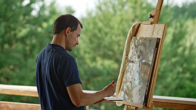 A male artist paints a picture on an easel on the street. Art as a hobby in nature. High quality 4k footage