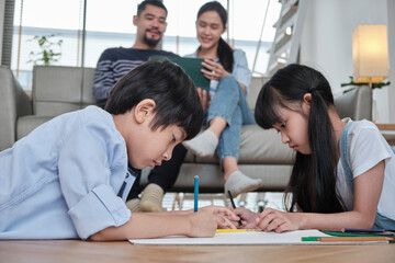 Asian Thai siblings are lying on living room floor, drawing homework with colored pencils together, parents leisurely relax on a sofa, lovely happy weekend activity, and domestic wellbeing lifestyle.