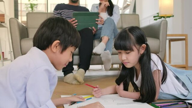 Asian Thai siblings are lying on living room floor, drawing homework with colored pencils together, parents leisurely relax on a sofa, lovely happy weekend activity, and domestic wellbeing lifestyle.