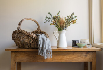 Wicker basket with grey scarf, Australian native flowers in white jug and assorted bowls on oak...