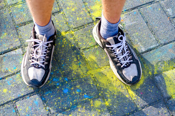 Men's feet in sneakers in selective focus stand on asphalt painted with Holi colors. Holi...
