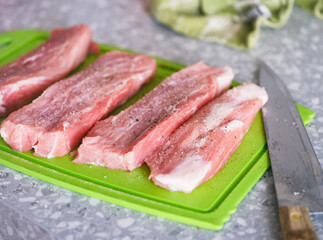 Peppered and salted raw pork slices on a green cutting board and knife on a kitchen table