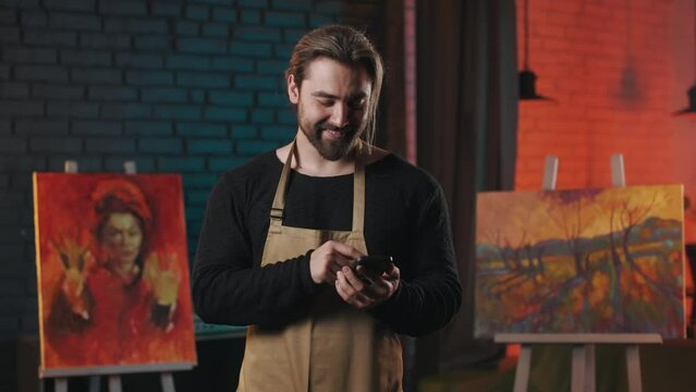 Caucasian art master holding modern smartphone and scrolling news in social network. Handsome male painter taking break from creative process with cell phone in hands.