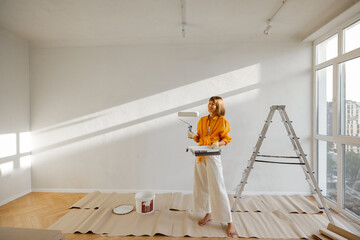 Portrait of a young woman making repairing in apartment, standing with paint roller near ladder in...