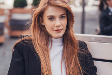 Close-up portrait of an elegant young caucasian woman looking convincingly at the camera while sitting on the park bench of the office building. Surfing the