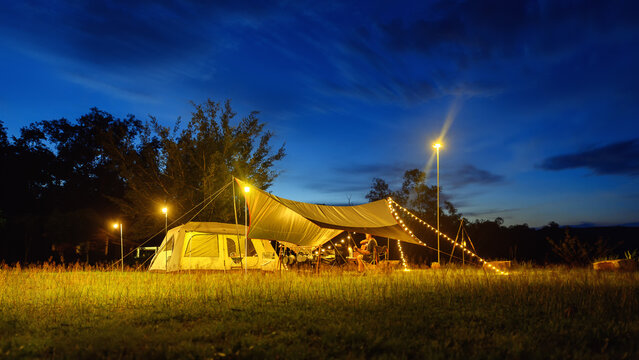 Outdoor camping tent with tarp or flysheet on grass courtyard and warm night light under dark blue sky twilight time, family vacation picnic on holiday relax, Overview of camping of family tourist.