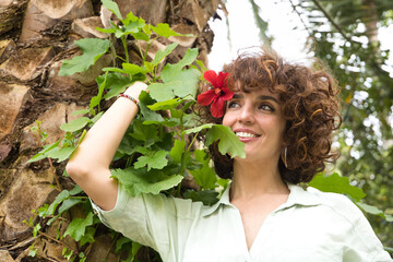 Portrait of mature and attractive woman with curly brown hair and a red flower in her ear, smiling and dreamy look, leaning on a palm tree with green leaves. Spring concept, flowers, hairstyle, curls.