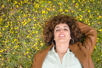Portrait of attractive mature woman with curly brown hair and leather jacket lying on the lawn surrounded by yellow flowers. Spring concept, flowers, beauty, hairstyle, curls.