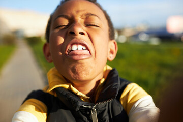 Funny self-portrait of african american boy kid grimacing, making faces playing with smartphone camera taking selfie outdoor while walking in park. Children and technology, gadgets and childhood - obrazy, fototapety, plakaty