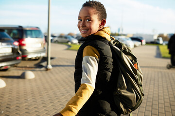 Back view of happy smiling african american schoolboy with backpack on shoulders walking down city street, going to school or returning from extra classes, sport center on sunny spring day