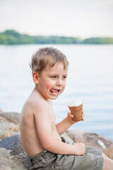 A cute blond boy appetizingly eats ice cream in the summer, sitting on the bank of the river. Cool off by the water. Funny facial expression.