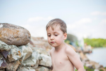 A cute blond boy experiences the joy of walking on a summer day by the river, sitting on the juicy grass. Funny facial expression. summer heat, a child without a T-shirt.