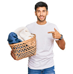 Young handsome man holding laundry basket pointing finger to one self smiling happy and proud
