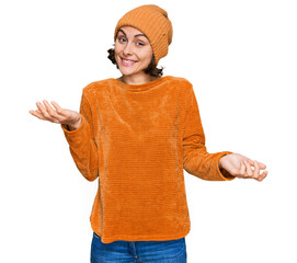 Young hispanic woman wearing casual clothes and wool cap clueless and confused expression with arms...