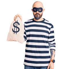 Young handsome man wearing burglar mask holding money bag with dollar symbol thinking attitude and...
