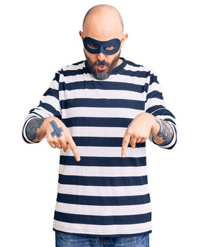 Young handsome man wearing burglar mask pointing down with fingers showing advertisement, surprised face and open mouth