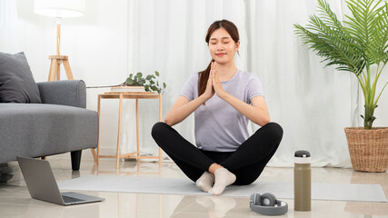Fototapeta na wymiar Young woman is relaxing into the lotus position for meditation on mat while taking yoga online class