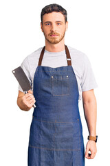 Handsome young man with bear wearing professional apron holding knife thinking attitude and sober...