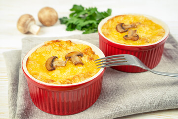 Chicken and mushroom casserole with golden crust, julienne with baked cheese, creamy gratin in portion ramekin clay pot on white wooden background. French cuisine dish, close up