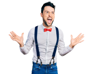 Hispanic man with beard wearing hipster look with bow tie and suspenders crazy and mad shouting and yelling with aggressive expression and arms raised. frustration concept.