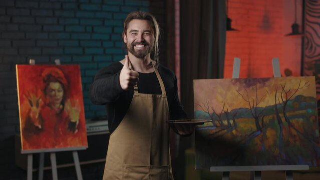 Happy bearded man standing near beautiful drawing and showing thumb up gesture. Caucasian art master in apron satisfied and proud of his masterpiece.
