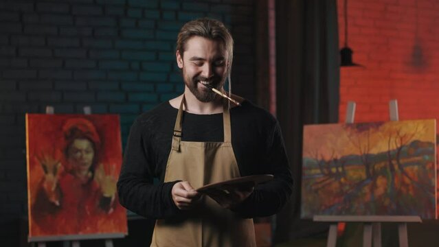 Positive caucasian artist with paintbrush in mouth and color palette in hands shrugging with shoulders while looking at camera at art studio. Favorite creative hobby.