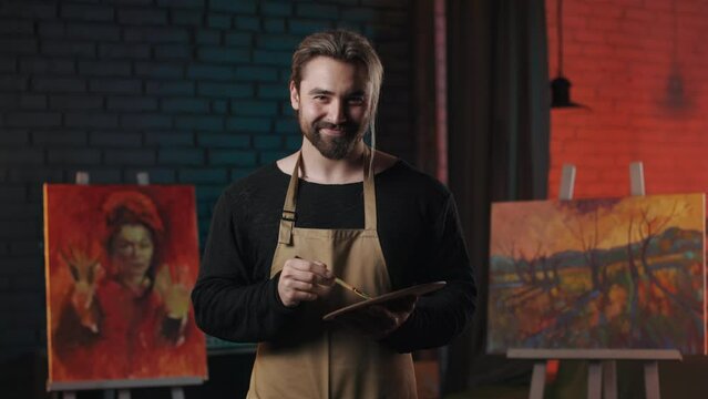 Happy bearded man in protective brown apron blending various colors on art palette with paintbrush. Talented male painter spending free time for favorite creative hobby.