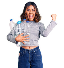 Young beautiful mixed race woman holding recycling plastic bottles screaming proud, celebrating victory and success very excited with raised arms