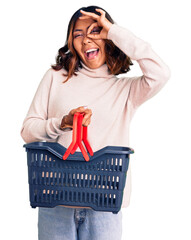 Young beautiful mixed race woman holding supermarket shopping basket smiling happy doing ok sign with hand on eye looking through fingers