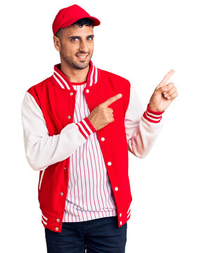 Young hispanic man wearing baseball uniform smiling and looking at the camera pointing with two hands and fingers to the side.