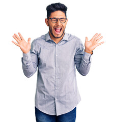 Fototapeta premium Handsome latin american young man wearing business clothes and glasses celebrating mad and crazy for success with arms raised and closed eyes screaming excited. winner concept