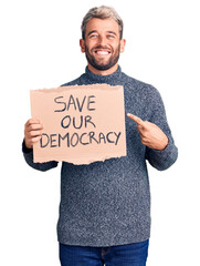 Young blond man holding save our democracy cardboard banner smiling happy pointing with hand and finger