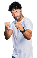 Hispanic young man with beard wearing casual white t shirt ready to fight with fist defense gesture, angry and upset face, afraid of problem