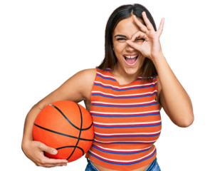 Young brunette woman holding basketball ball smiling happy doing ok sign with hand on eye looking through fingers