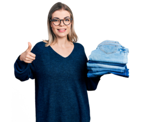 Young blonde woman holding stack of folded jeans smiling happy and positive, thumb up doing excellent and approval sign