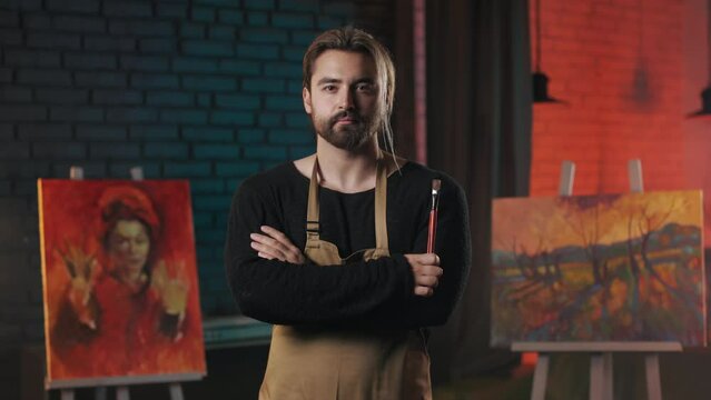 Confident male artist with paintbrush in hands standing in modern art space and looking at camera with calm face. Concept of people, creativity and inspiration.