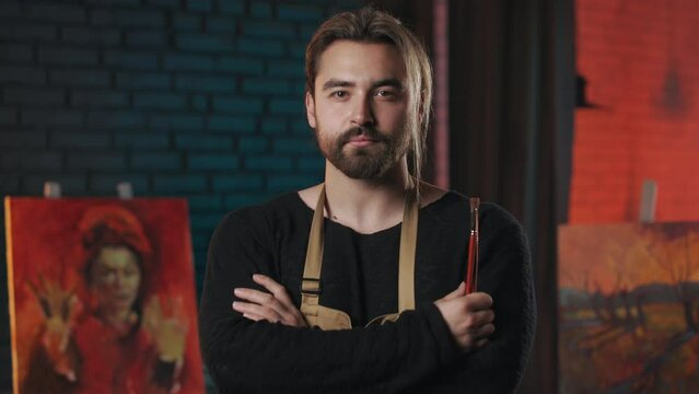 Caucasian young man with neutral facial expression standing at workshop and keeping arms crossed. Male painter with stylish beard and haircut wearing protective apron at work.