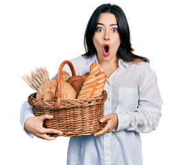 Beautiful hispanic woman holding wicker basket with bread scared and amazed with open mouth for surprise, disbelief face