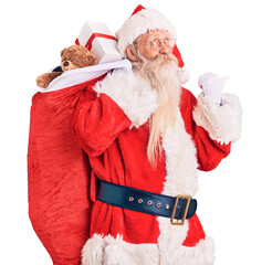 Old senior man with grey hair and long beard wearing santa claus costume holding bag with presents pointing thumb up to the side smiling happy with open mouth