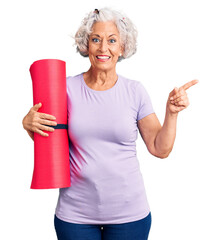 Senior grey-haired woman holding yoga mat smiling happy pointing with hand and finger to the side