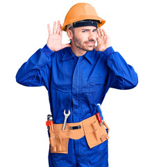 Young hispanic man wearing worker uniform trying to hear both hands on ear gesture, curious for gossip. hearing problem, deaf