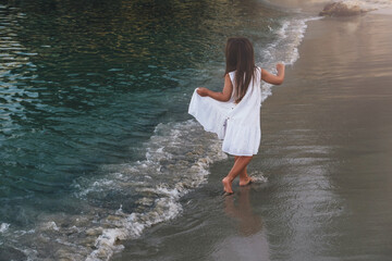 A little girl in a white dress walks along the line of the sea. - obrazy, fototapety, plakaty