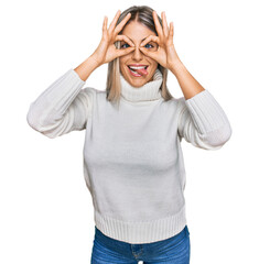 Beautiful blonde woman wearing casual turtleneck sweater doing ok gesture like binoculars sticking tongue out, eyes looking through fingers. crazy expression.