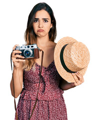 Beautiful hispanic woman holding vintage camera and summer hat depressed and worry for distress,...