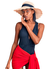 Young african american woman wearing swimsuit and summer hat hand on mouth telling secret rumor, whispering malicious talk conversation