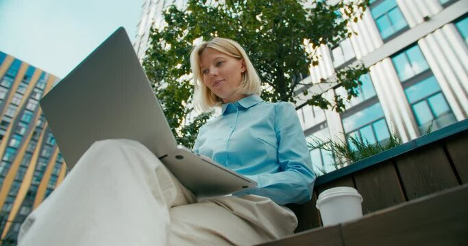 Young woman university student learning online using laptop computer, taking notes, watching webinar or virtual education remote class studying outdoor sitting outside uni campus area.