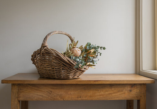 Wicker basket with Australia native flowers and eucalyptus leaves on oak side table against beige wall