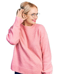 Young blonde woman wearing casual clothes and glasses smiling with hand over ear listening an hearing to rumor or gossip. deafness concept.